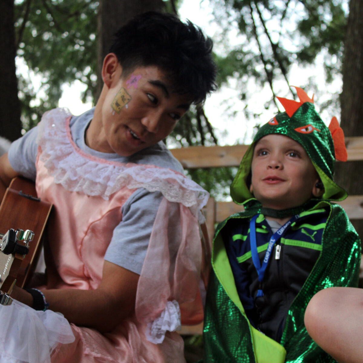 a camper in a dinosaur costume sits on the stage and sings as a camp counselor plays guitar
